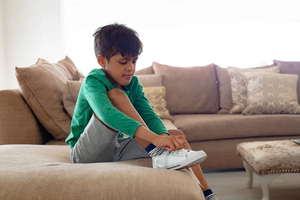 快適な家のリビングルームでソファに靴ひもを結ぶかわいいアメリカの男の子の側面図 — ストック写真