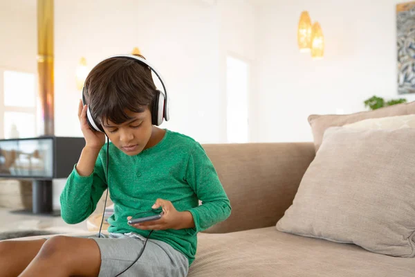 美国男孩前视图与耳机在舒适的家庭在手机上听音乐 — 图库照片
