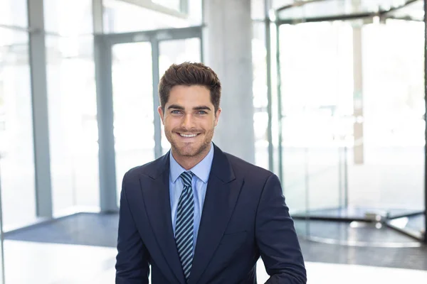 現代のオフィスに立ちながらカメラに微笑む若い白人ビジネスマンの正面図 — ストック写真