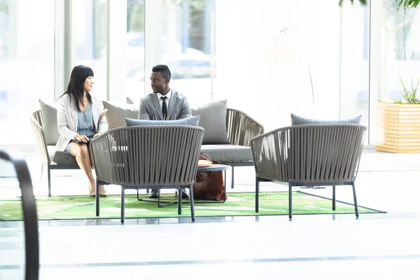 アジアの若い女性とアフリカ系アメリカ人の男性幹部が現代のオフィスでベンチで話す正面図 — ストック写真
