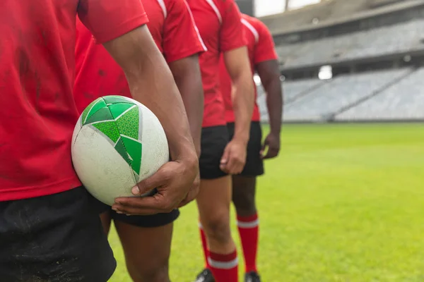 Mid section close up of diverse male rugby players standing together with rugby ball in stadium.