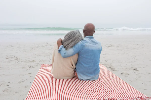 坐在海滩上的红色和白色条纹野餐毯上的快乐老年夫妇的后视图 正宗的退休老人生活理念 — 图库照片