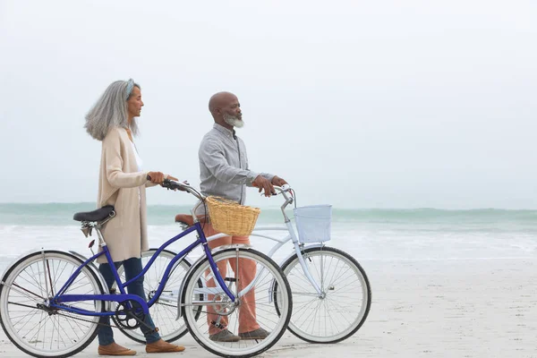 在美丽的一天 英俊的非洲裔美国夫妇骑着自行车在沙滩上行走 双手插在口袋里 正宗的退休老人生活理念 — 图库照片