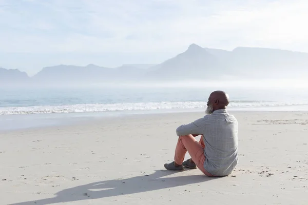 英俊的非洲裔老人坐在沙滩上 在美丽的一天看着沙滩上的大海和山脉的后视图 正宗的退休老人生活理念 — 图库照片