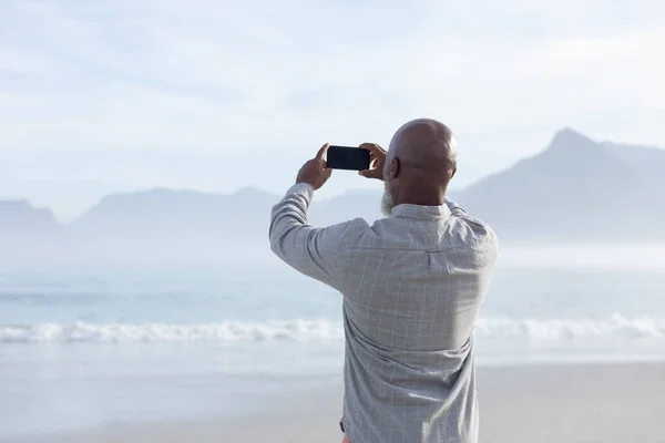 美しい日にビーチで海と山の写真を撮るハンサムなアフリカ系アメリカ人の男性の背面図 本物のシニアリタイアライフコンセプト — ストック写真