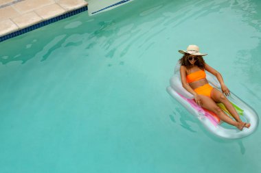 High angle view of mixed-race woman in bikini relaxing on a inflatable tube in swimming pool at the backyard of home. clipart