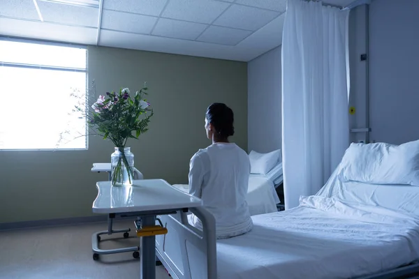 病棟のベッドに座りながら目をそらす混合人種女性患者の後方図 — ストック写真