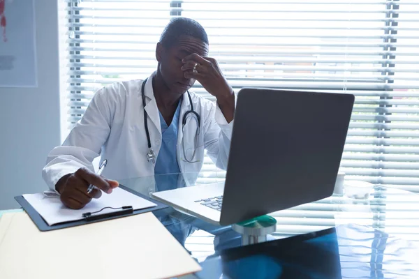 心配したアフリカ系アメリカ人男性医師が病院の机のクリップボードに書き込みながら目をこする正面図 — ストック写真