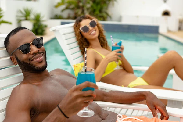 在游泳池附近的日光浴躺椅上放松时 幸福的多元化夫妇享用鸡尾酒的前视图 — 图库照片