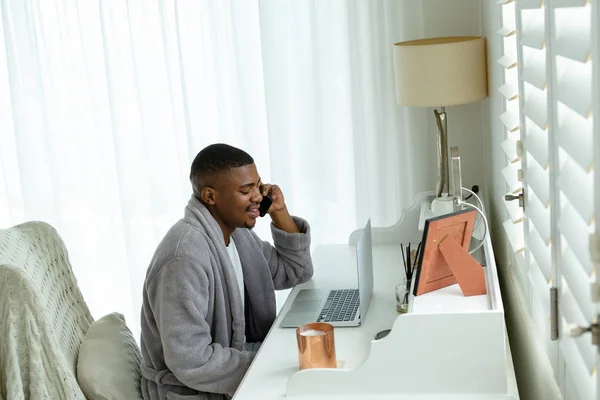 快適な家の寝室の机でラップトップで作業しながら 携帯電話で話しているアフリカ系アメリカ人の男性の側面図 — ストック写真