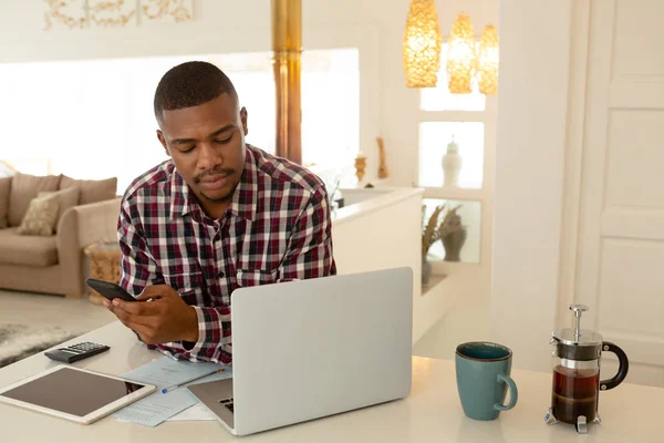 快適な家のテーブルで携帯電話とラップトップを使用してアフリカ系アメリカ人の男性の正面図 — ストック写真