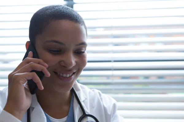 病院で携帯電話で話す首の周りに聴診器を持つ幸せな混合レースの女性の肖像画 — ストック写真