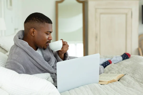 Side view of African-american man drinking coffee while using laptop on bed in bedroom at comfortable home.