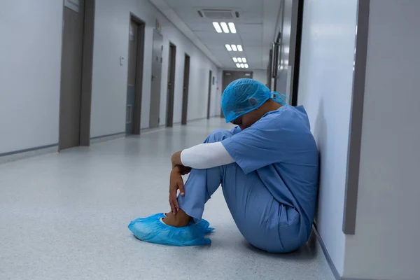 紧张混合种族女外科医生坐在地板上 眼睛闭上在医院走廊的侧视图 — 图库照片