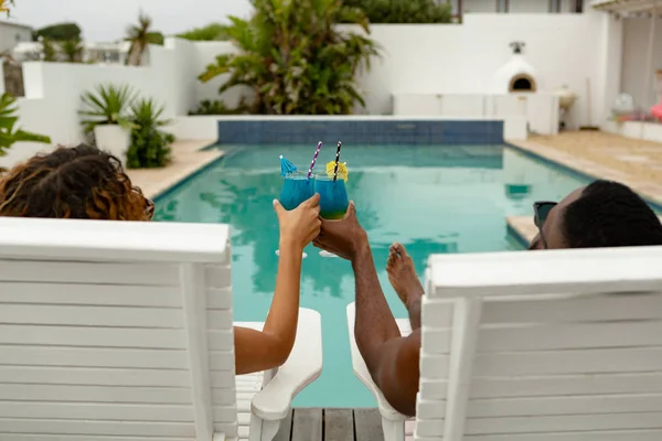 可爱的多样化夫妇在后院游泳池附近的太阳椅上放松时 一边举杯鸡尾酒的后视图 — 图库照片
