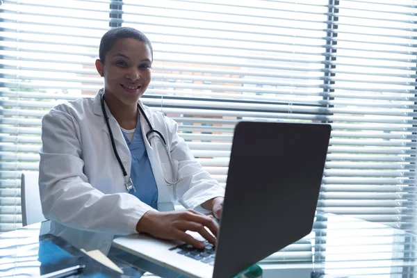 病院の机でカメラを見ながらノートパソコンを使って首の周りに聴診器を持つ混合人種の女性医師の肖像 — ストック写真