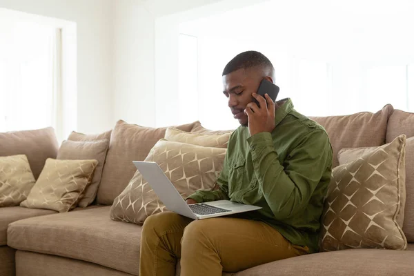 快適な家のリビングルームでソファでラップトップを使用しながら 携帯電話で話す気配りのアフリカ系アメリカ人男性の側面図 — ストック写真