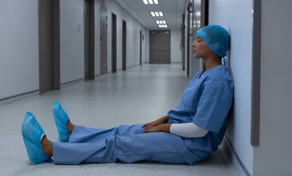 紧张混合种族女外科医生坐在医院走廊上闭着眼睛的侧视图 — 图库照片