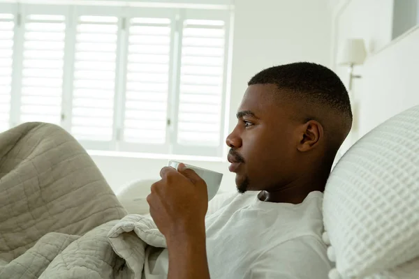 快適な家の寝室でベッドでコーヒーを飲む思慮深いアフリカ系アメリカ人男性の側面図 — ストック写真