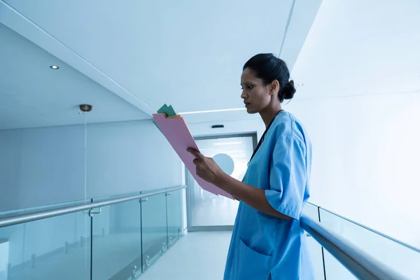 病院の廊下で医療報告書を見ている混合人種女性外科医の側面図 — ストック写真