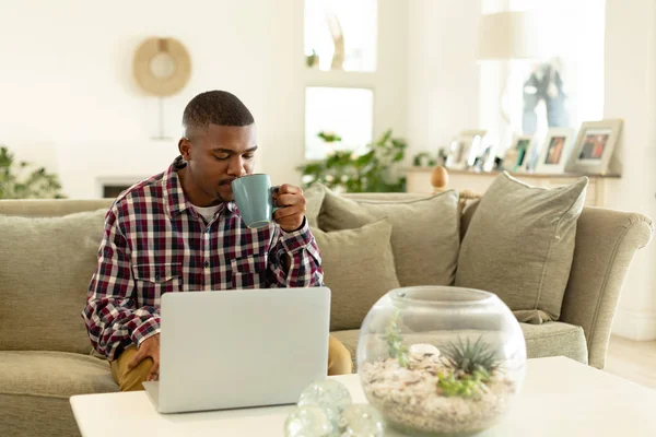 快適な家のリビングルームでソファでラップトップを使用しながらコーヒーを飲むアフリカ系アメリカ人の男性の正面図 — ストック写真