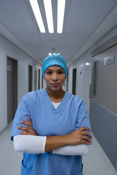 病院の廊下でカメラを見ながら腕を組んで立っている混合人種女性外科医の肖像 — ストック写真