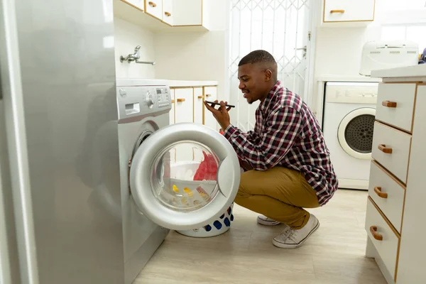 自宅の台所の洗濯機に衣類を入れながら携帯電話で話すアフリカ系アメリカ人男性の側面図 — ストック写真