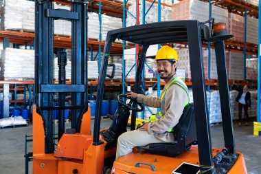 Front view of happy mature Asian male worker sitting in forklift and looking at camera in warehouse. clipart