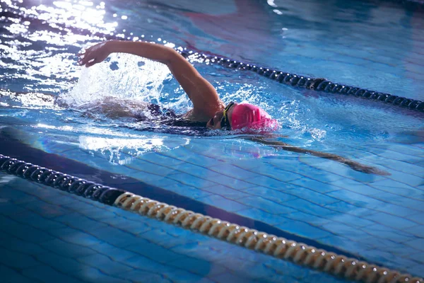 一位非洲裔美国妇女戴着粉红色泳帽 戴着护目镜在游泳池里做自由泳的侧视图 — 图库照片