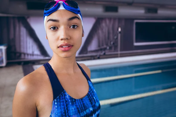 一位年轻的非洲裔美国妇女穿着泳衣 戴着泳帽 戴着护目镜 站在体育场内的奥林匹克大小的游泳池旁 看着镜头 — 图库照片