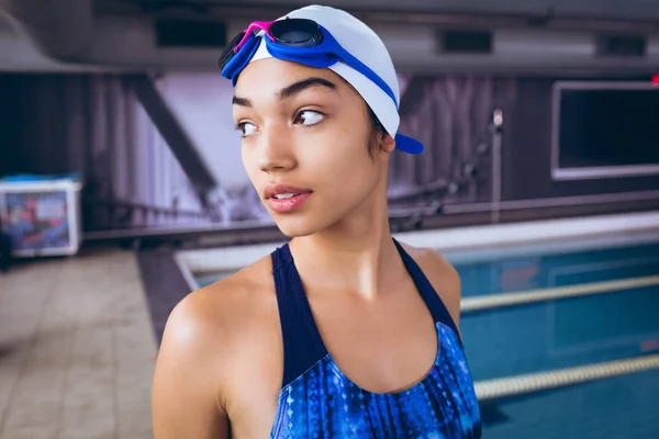 一位年轻的非洲裔美国妇女穿着泳衣 戴着泳帽 戴着护目镜 站在体育场内的奥林匹克大小的游泳池旁 — 图库照片