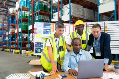Front view of mature African-american male supervisor with his diverse coworkers discussing over laptop at desk in warehouse.  clipart