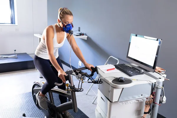 在体育中心房间内骑自行车时 一名白种运动妇女使用连接到监视器的面具进行体能测试的侧视图 — 图库照片