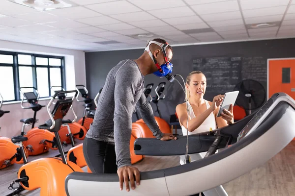 Side view of an African-American athletic man doing a fitness test using a mask while using a treadmill and a Caucasian woman showing his results inside a room at a sports center.