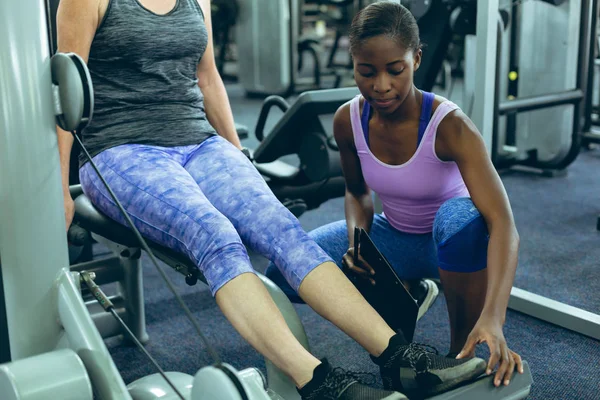 年轻适合非洲裔美国女教练的前视图 协助活跃妇女锻炼在健身中心的腿卷曲机 — 图库照片