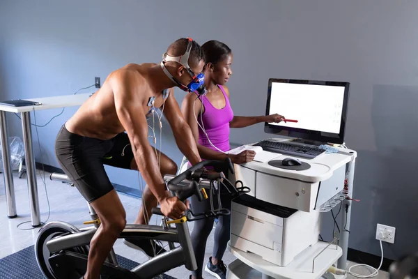 一名赤裸裸的非裔美国运动男子在骑自行车时 用与显示器相连的面具进行体能测试 一名非洲裔美国妇女向体育中心房间内的监视器显示结果 — 图库照片