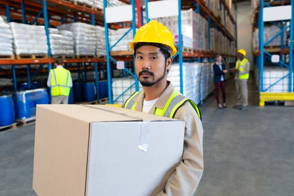 英俊的亚洲男性工人携带纸板箱和在仓库看相机的正面视图 — 图库照片