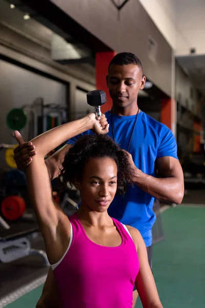 ダンベルを持つアフリカ系アメリカ人女性と スポーツセンターの部屋の中で彼女を手伝うアフリカ系アメリカ人の男性の正面図 — ストック写真