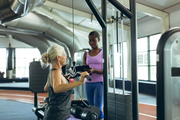 美丽的非洲裔美国女教练侧视图协助活跃的高级高加索妇女在健身工作室的拉拔机 — 图库照片
