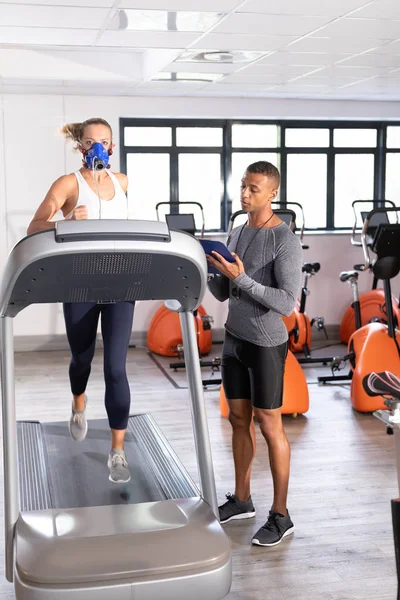 Front view of a Caucasian athletic woman doing a fitness test using a mask while using a treadmill and an African-American man monitoring her inside a room at a sports center.