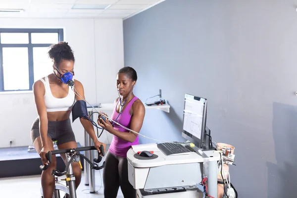 エクササイズバイクに乗りながらモニターに接続されたマスクを使用してフィットネステストを行うアフリカ系アメリカ人のアスレチック女性と スポーツセンターの部屋の中で血圧を測定するアフリカ系アメリカ人女性の正面図 — ストック写真