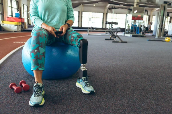 フィットネスセンターでエクササイズボールに座っている間 携帯電話を使用して脚切断患者を持つ障害のあるアクティブな女性の低いセクション — ストック写真
