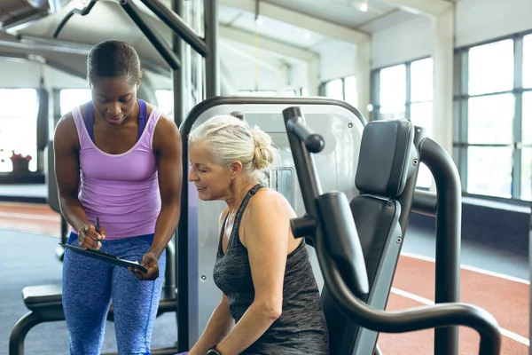 前视图的非洲裔美国女教练讨论剪贴板与活跃的高级高加索妇女在健身工作室 — 图库照片