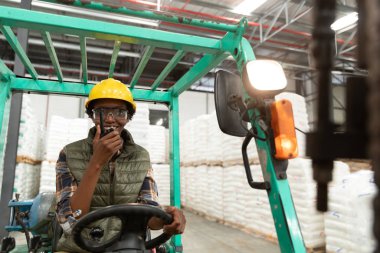 Happy female worker talking on walkie-talkie while driving forklift in warehouse. clipart