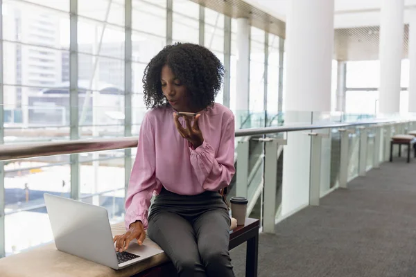 坐在现代办公室大厅里 年轻的非洲裔女商人在笔记本电脑上工作 用手机交谈 — 图库照片