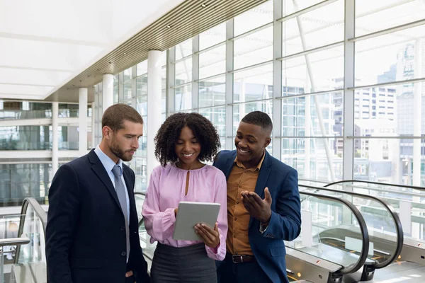 Front view of an African American and a Caucasian businessmen standing in the lobby of a modern office with an African American businesswoman looking at a tablet computer and talking.