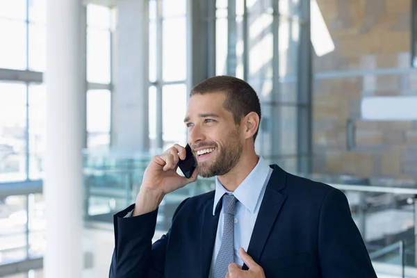 近代的なビジネスロビーのアトリウムで自分のスマートフォンで話している笑顔の若い白人ビジネスマンの正面図 — ストック写真