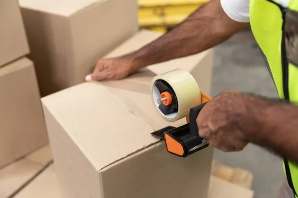 Close-up of male worker packing cardboard box with tape gun dispenser in warehouse.