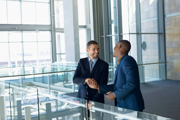 若い白人ビジネスマンと若いアフリカ系アメリカ人ビジネスマンが近代的なオフィスビルに立って握手する3四半期の長さの眺め — ストック写真