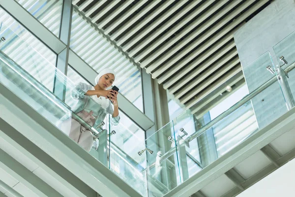 現代のオフィスの廊下で手すりに寄りかかって携帯電話を使用するヒジャーブのビジネスウーマンの低角度ビュー — ストック写真